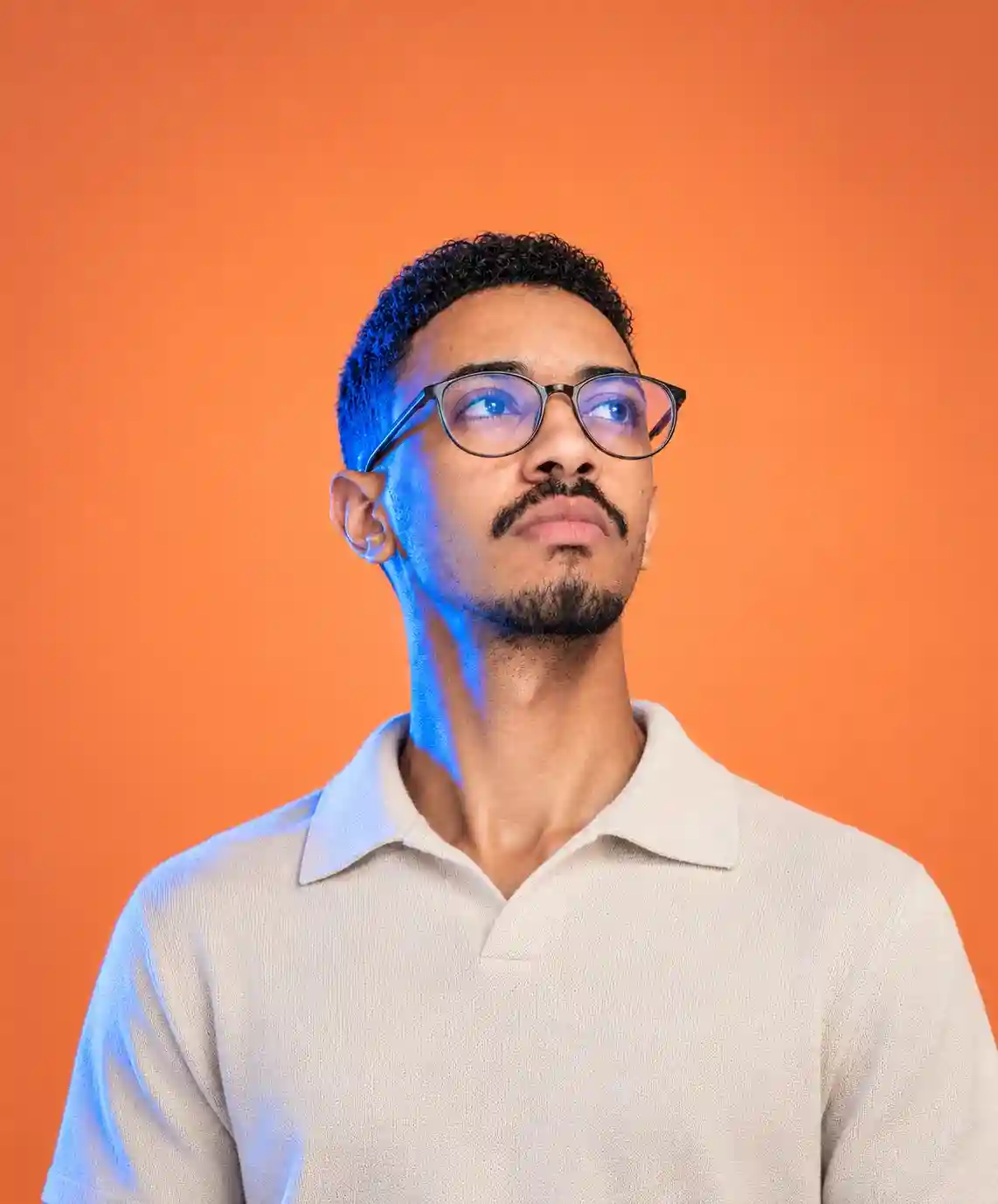 Young man with glasses and a mustache looking thoughtfully upward against an orange background